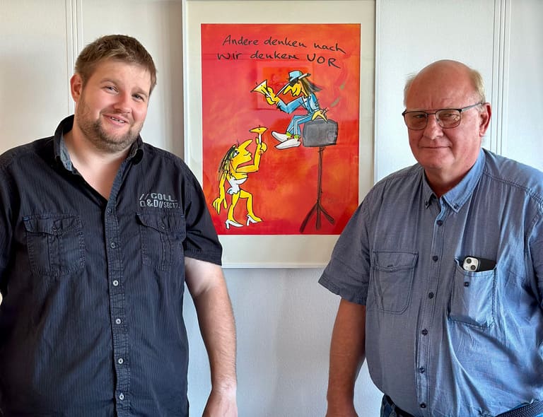 Eckhard und Alex Wendt von KLH Kabel- und Leitungsbau GmbH lächeln im Büro neben einem kreativen Cartoon-Poster zum Thema "Vorausdenken".